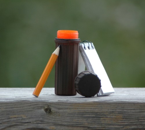 A pencil, a notepad, and a container are arranged on a wooden surface with a blurred green background.