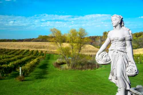 A white statue of a woman holding baskets stands in a vineyard with rolling hills and a blue sky in the background.