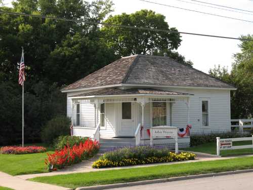 A small white house with a porch, surrounded by colorful flowers and greenery, featuring an American flag.