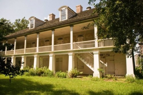 A historic two-story house with a porch, surrounded by greenery and trees, under a clear blue sky.