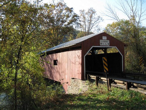A red covered bridge surrounded by trees, with a sign indicating clearance height, over a small stream.