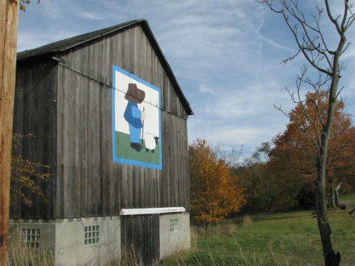 A wooden barn with a painted mural of a person holding a sheep, surrounded by autumn trees and a clear sky.
