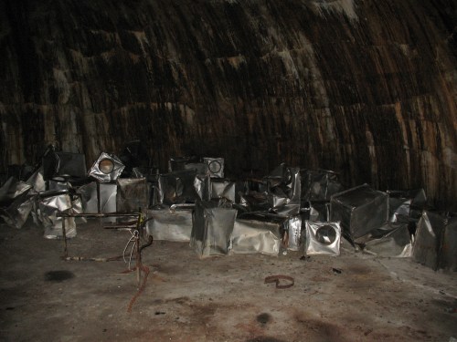 A dimly lit, abandoned space with scattered metallic containers and a rough, textured wall in the background.