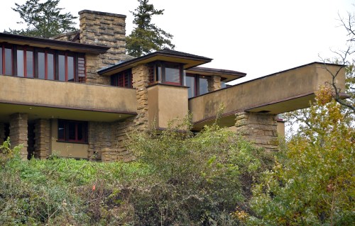 A modernist house with stone and stucco exterior, featuring large windows and a cantilevered balcony, surrounded by greenery.