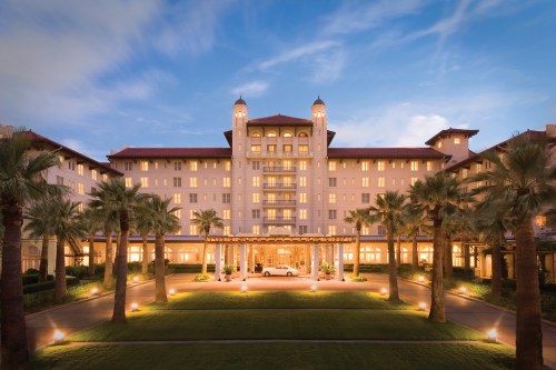 A grand hotel with palm trees in front, illuminated at dusk, showcasing elegant architecture and a welcoming entrance.