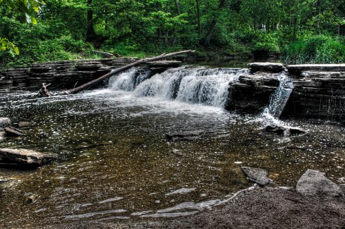 A serene waterfall cascades over rocky ledges into a calm pool, surrounded by lush green trees and foliage.