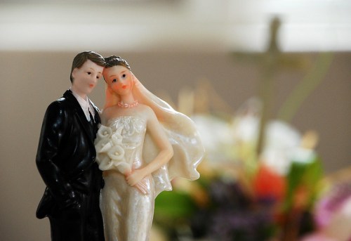 A close-up of a wedding cake topper featuring a bride and groom, with a blurred background of flowers and a cross.