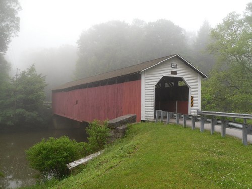A red covered bridge shrouded in fog, surrounded by green trees and a calm waterway.