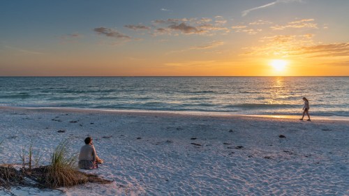 A serene beach at sunset, with one person sitting on the sand and another walking along the shoreline.