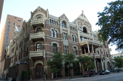 Historic brick building with ornate architecture, balconies, and surrounding greenery, set against a city backdrop.