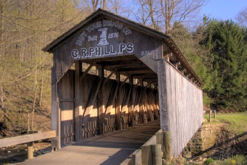 A historic wooden covered bridge with a sign reading "C.R. Phillips, Built 1876," surrounded by trees and a clear sky.