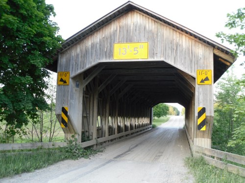 A wooden covered bridge with height clearance signs, leading to a dirt road surrounded by greenery.