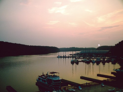 Sunset over a calm marina with boats docked, surrounded by trees and a hazy sky.