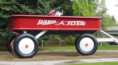 A large red Radio Flyer wagon with two people waving from the top, set in a park with trees in the background.