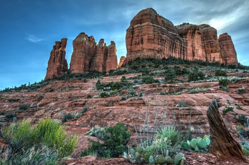 Red rock formations rise against a blue sky, surrounded by desert vegetation and cacti in a rugged landscape.