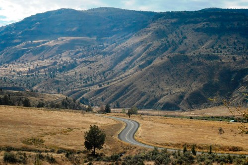 A winding road cuts through a dry, hilly landscape with sparse vegetation under a cloudy sky.