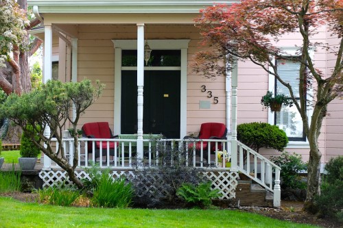 A charming house with a pink exterior, featuring a porch with two red chairs and lush greenery surrounding it.