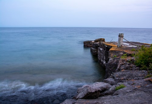 A rocky shoreline meets calm, blue waters under a cloudy sky, with a wooden post and greenery in the foreground.