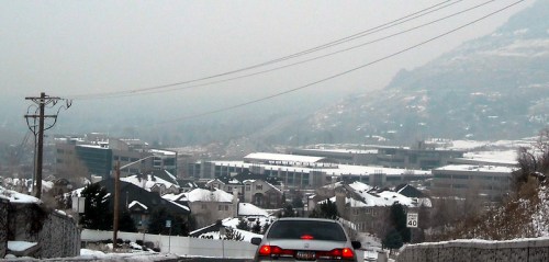 A car drives down a snowy road with a view of buildings in the distance, shrouded in fog. Power lines are visible.