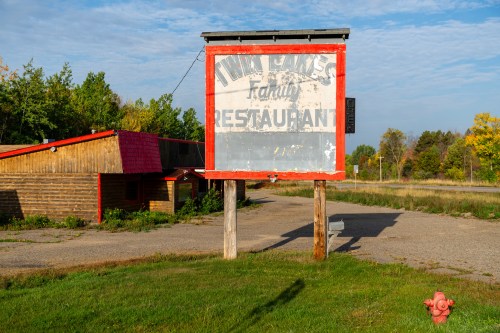 A weathered sign for "Twin Gables Family Restaurant" stands near a rustic building, surrounded by greenery and a clear sky.