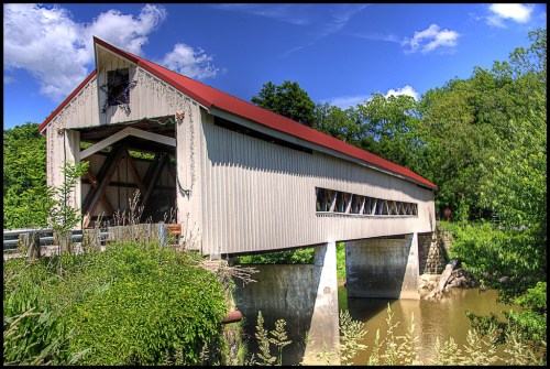 A scenic view of a covered bridge over a calm river, surrounded by lush greenery and a clear blue sky.