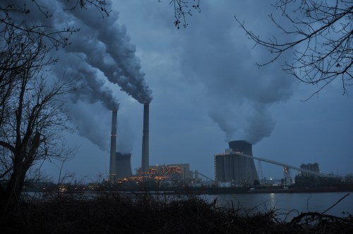 A power plant with tall smokestacks emitting smoke, set against a dark, cloudy sky near a river.