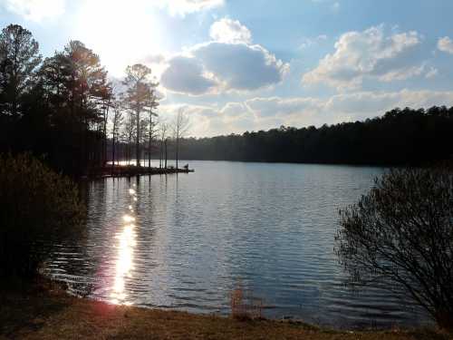 A serene lake scene with sunlight reflecting on the water, surrounded by trees and a cloudy sky.