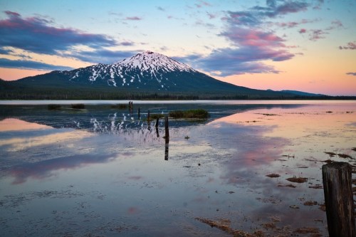 A serene lake reflects a snow-capped mountain at sunset, with colorful clouds and wooden posts in the foreground.