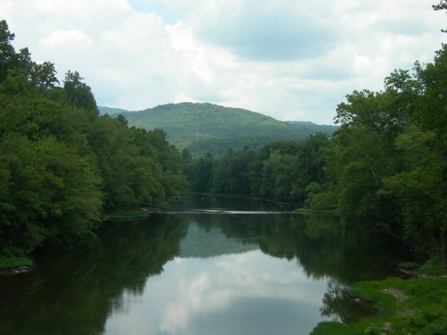 A serene river reflects lush green trees and distant mountains under a cloudy sky.