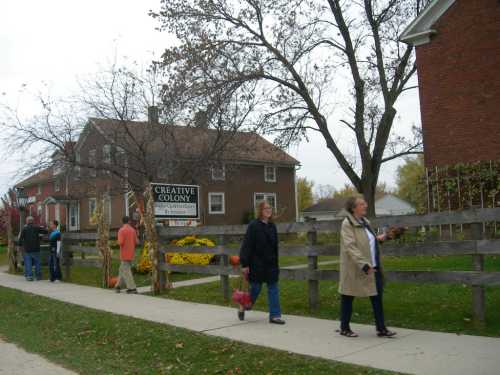 People walking along a sidewalk past a house with a sign for "Creative Colony" and autumn decorations.