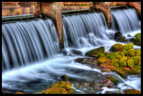A serene waterfall cascades over stone barriers, surrounded by lush green moss and smooth rocks.