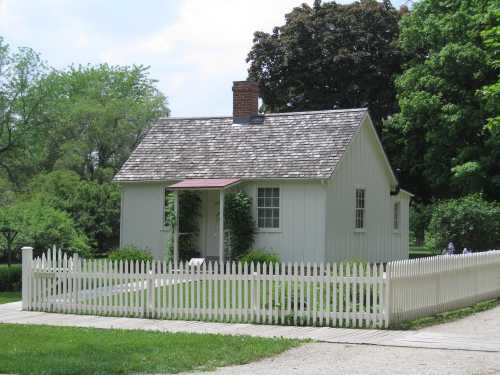 A small white house with a red roof, surrounded by greenery and a white picket fence on a sunny day.