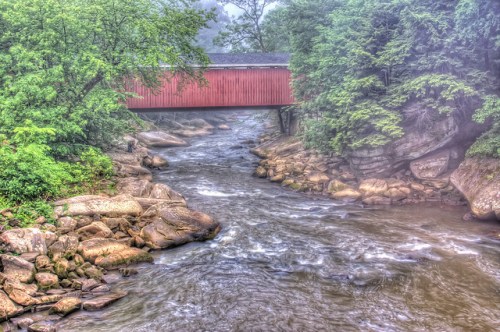 A red covered bridge spans a misty river, surrounded by lush greenery and rocky banks.