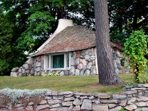A quaint stone cottage with a rounded roof, surrounded by trees and a stone wall, set in a lush green landscape.