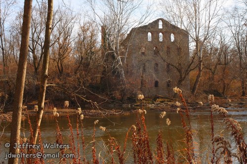 Ruins of a stone building beside a calm river, surrounded by bare trees and autumn foliage.