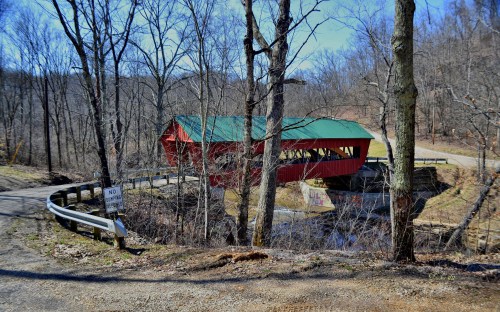 A red covered bridge with a green roof spans a small creek, surrounded by bare trees and a gravel road.