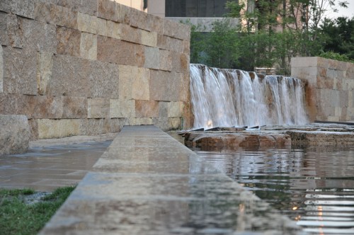 A serene waterfall cascades over stone, surrounded by greenery and reflecting in a calm pool of water.