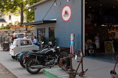 A vintage shop exterior with classic cars, a motorcycle, and a whimsical statue outside.