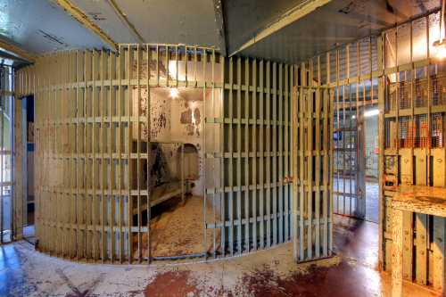 Interior of a prison cell block featuring barred doors and a circular cell area with peeling paint and dim lighting.