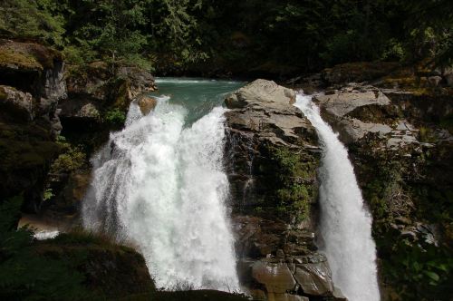 A scenic waterfall cascading over rocky cliffs, surrounded by lush green trees and a clear blue river.