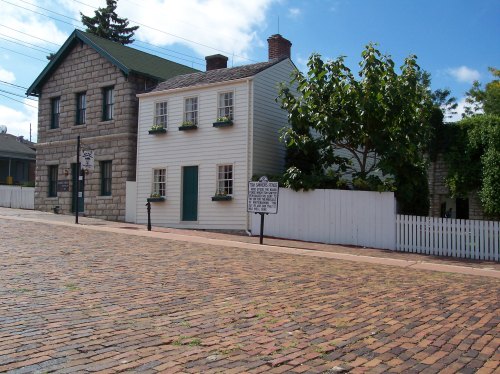 A quaint white house with green accents and flower boxes, set on a cobblestone street near a stone building.