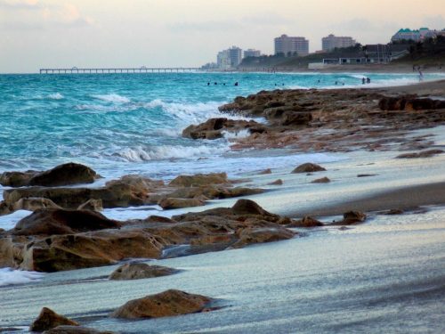 A serene beach scene with rocky shorelines, gentle waves, and distant buildings under a soft, cloudy sky.