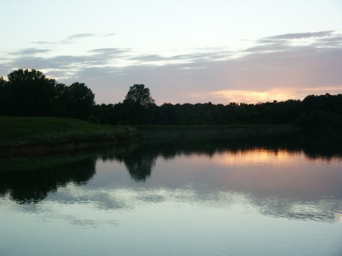 A serene lake at sunset, reflecting colorful clouds and surrounded by trees.