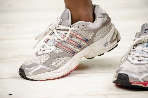 Close-up of a person's foot wearing gray and white Adidas running shoes with colorful accents on a light wooden surface.