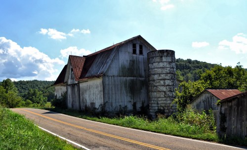 A weathered barn and silo beside a winding road, surrounded by lush greenery and hills under a blue sky.