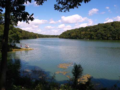 A serene lake surrounded by lush greenery and trees, with a few people on a small dock under a blue sky.