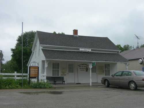 A white, single-story building with a gray roof, featuring a sign and a parked car in front. Overcast sky.