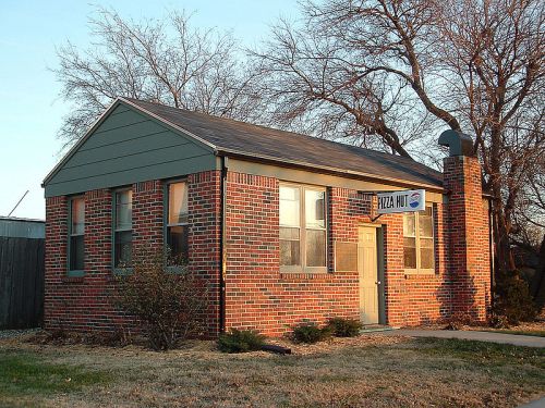 A small brick building with a gray roof and a sign reading "Pizza Auto," surrounded by trees and grass.