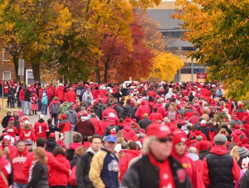 A large crowd of people in red clothing walking through a park with colorful autumn trees in the background.