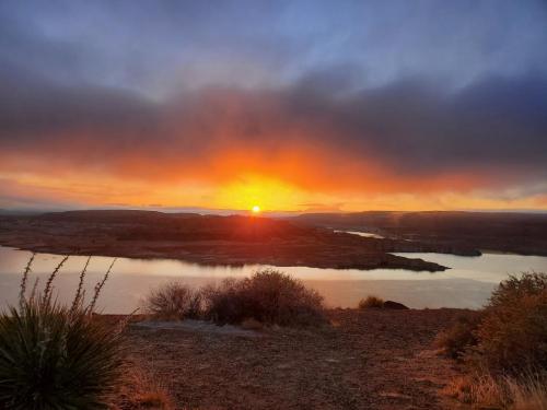 A vibrant sunset over a calm lake, with silhouettes of hills and shrubs in the foreground and clouds in the sky.